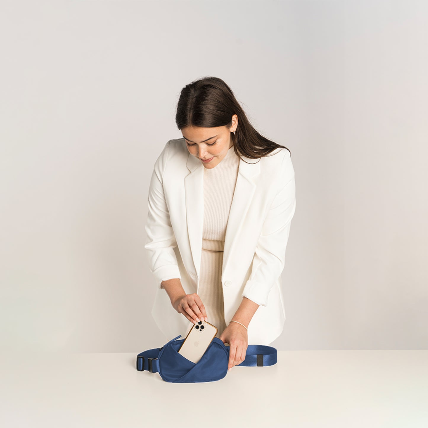 Woman in a white blazer holding a phone next to a Navy blue waist bag.