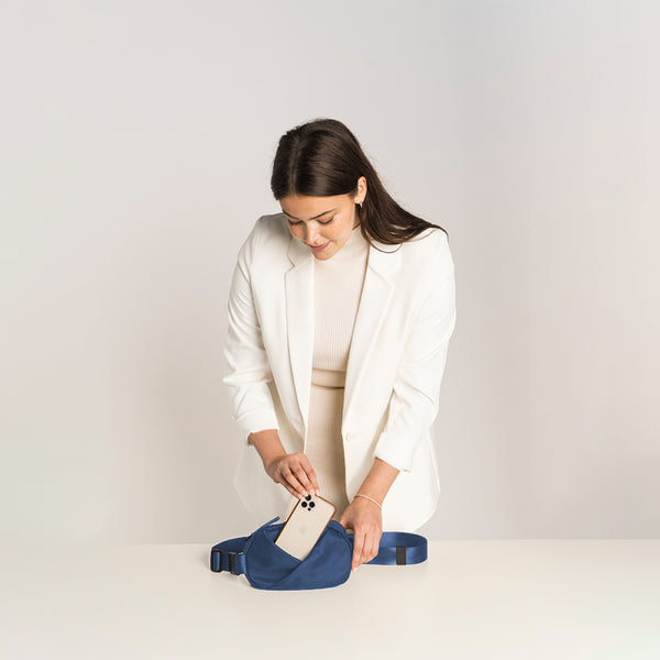 Woman in a white blazer holding a phone next to a Navy blue waist bag.