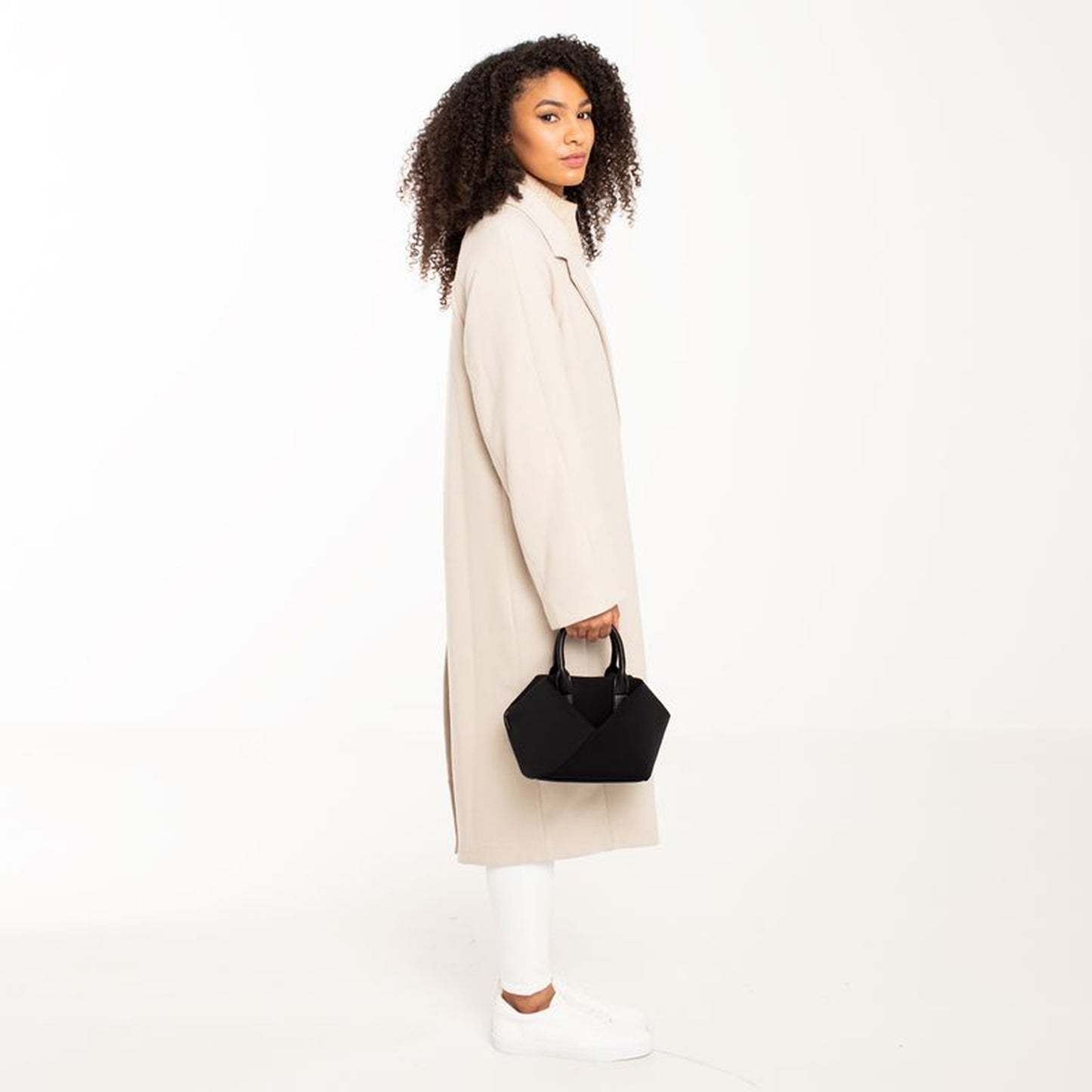 Woman wearing a long beige coat holding a Black + Gold Hardware & Black + Silver Hardware handbag.