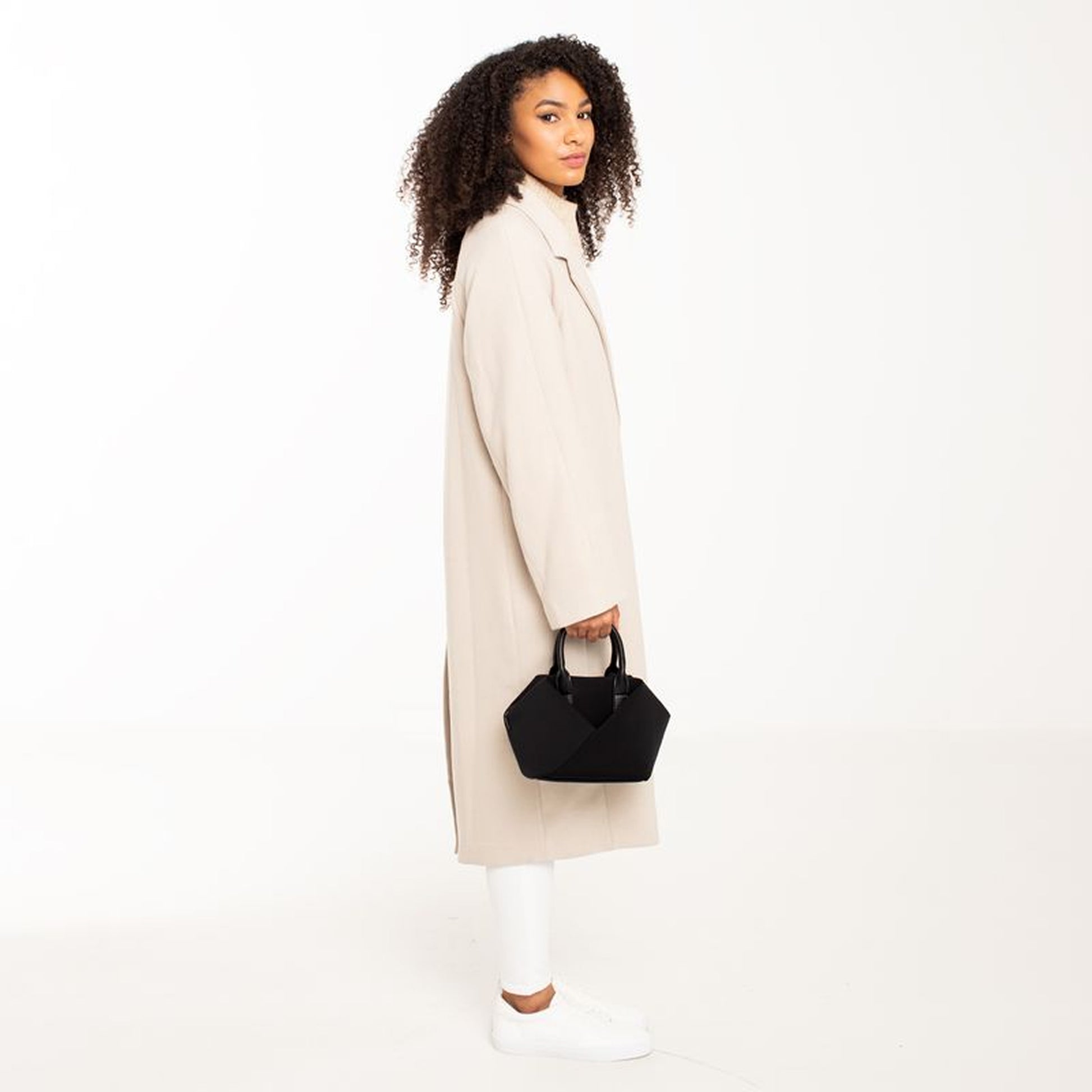 Woman wearing a long beige coat holding a Black + Gold Hardware & Black + Silver Hardware handbag.