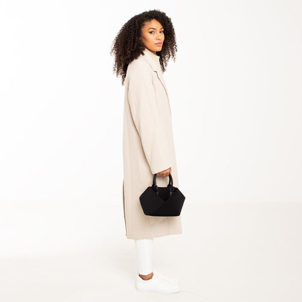 Woman wearing a long beige coat holding a Black + Gold Hardware & Black + Silver Hardware handbag.
