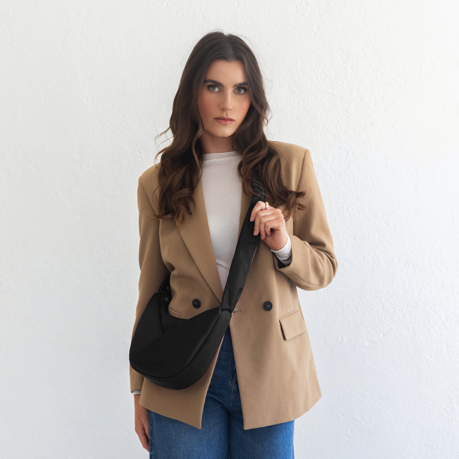 Woman wearing a beige blazer and blue jeans, holding a black bag against a white background