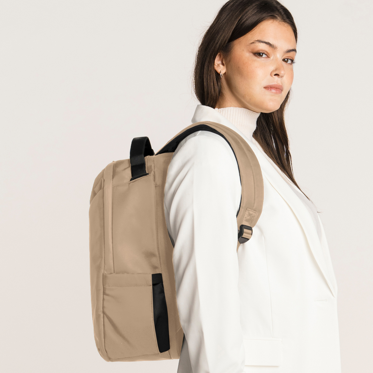  A woman wearing a white blazer and white turtleneck carries  Lux & Nyx Origami Backpack - 16" Laptop Backpack With Trolley Strap - 
Champagne Taupe