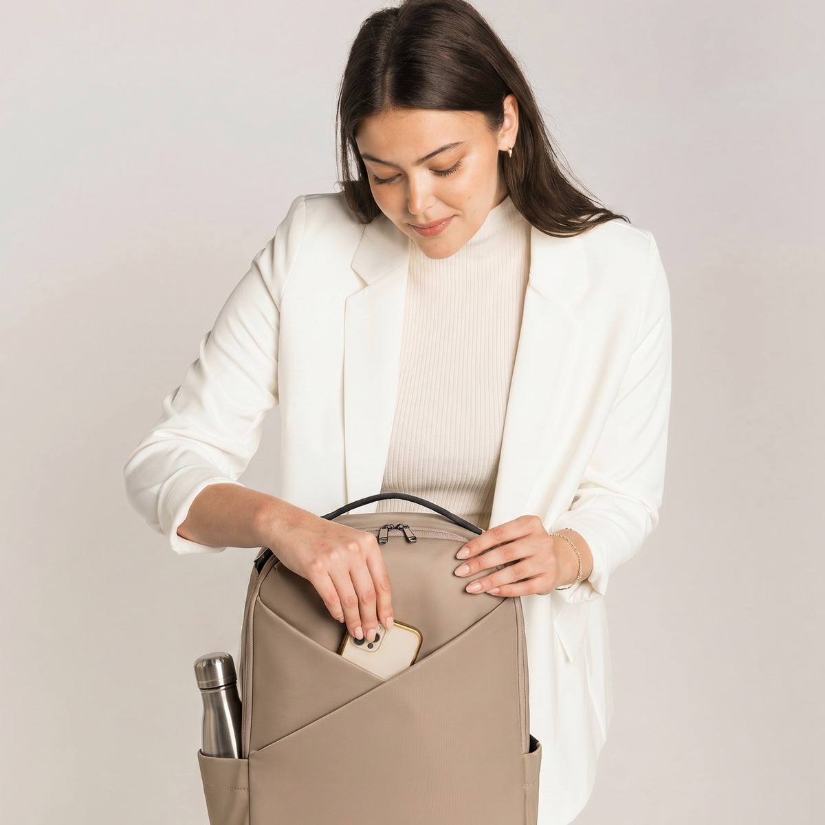 Lux & Nyx Origami Backpack - 16" Laptop Backpack With Trolley Strap - 
Champagne Taupe - A woman in a white blazer and cream ribbed top reaches into a beige backpack pocket, demonstrating storage capacity.