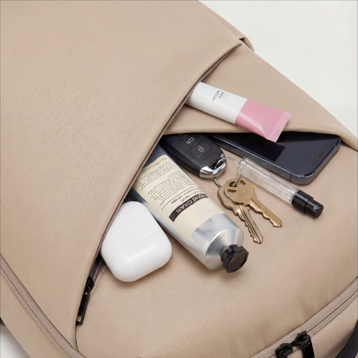 Lux & Nyx Origami Backpack - 16" Laptop Backpack With Trolley Strap - 
Champagne Taupe shown open with its front pocket unzipped, revealing everyday items inside: a smartphone, keys on a keychain, a small clear spray bottle, a white earbud case, a beige tube, and a cosmetic tube.