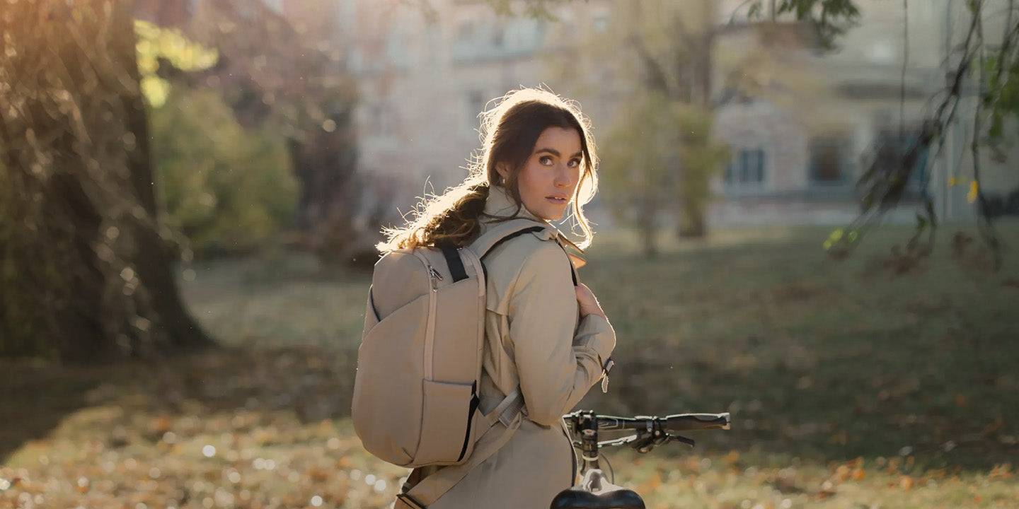 A woman wearing a beige jacket and carrying a large lux and nyx beige backpack stands outdoors in a sunlit park near a bicycle.