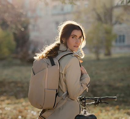 A woman wearing a lux and nyx beige backpack and matching beige trench coat stands outdoors next to a bicycle in a sunlit park setting.