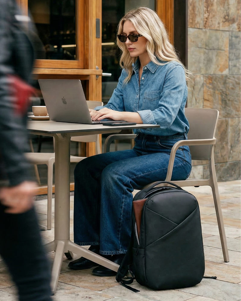 Woman using a laptop at an outdoor cafe with a black backpack next to her.