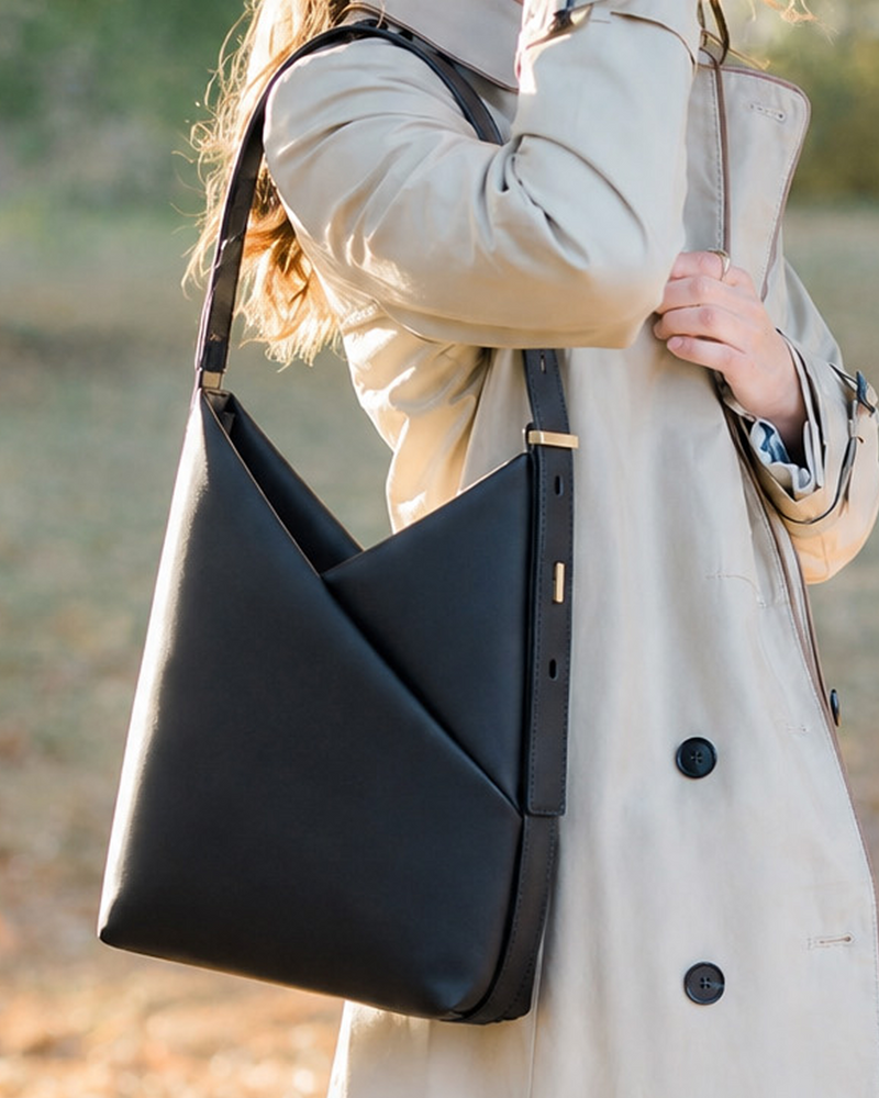 A woman wearing a beige trench coat carries a large black leather shoulder bag outdoors with a blurred natural background.