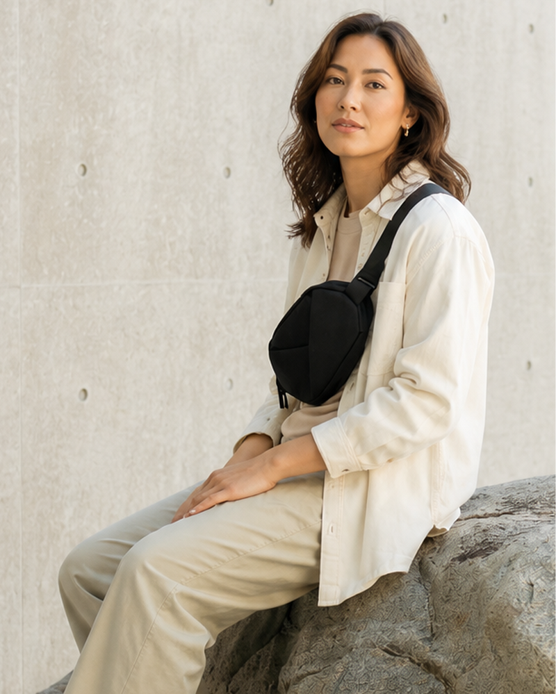 Woman sitting on a rock wearing a white shirt and beige pants, with a black bag over her shoulder against a light stone wall.