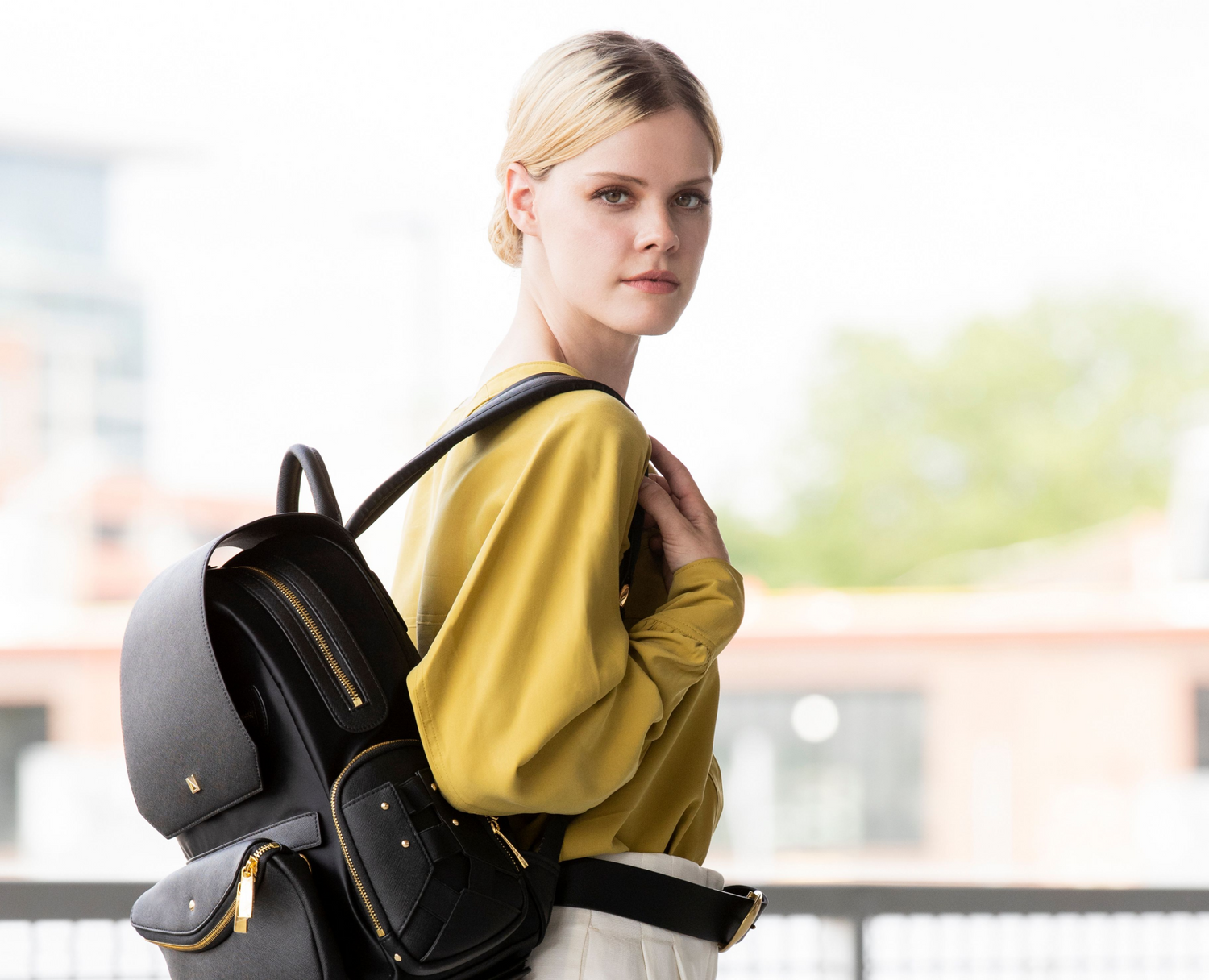 Black leather backpack with gold-tone hardware worn by a woman in a mustard blouse, featuring multiple zippered compartments.