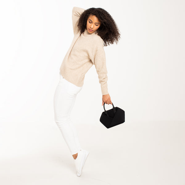 Woman in a beige sweater and white pants posing while holding a small black-with-gold-hardware / black-with-silver-hardware handbag against
